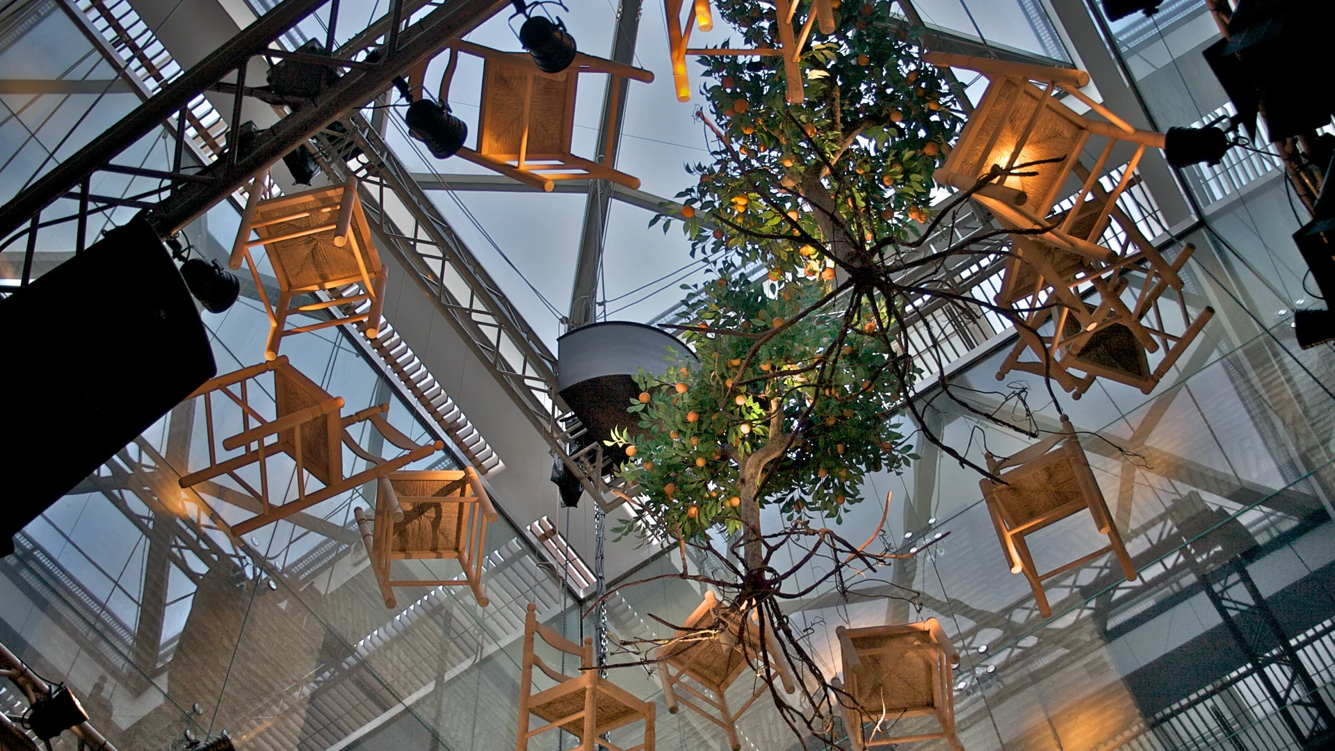 Looking up inside the museum's patio: wooden chairs with woven seats and an orange tree full of fruit hang in the air beneath a glass roof and stage lights