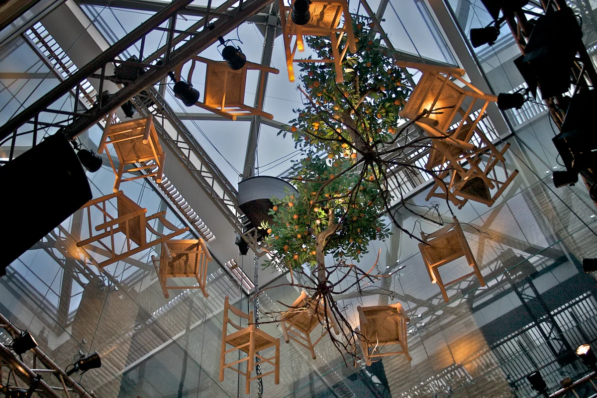 The patio seen from below: chairs with woven seats and an orange tree hang in the air under the glass roof, with stage lights around them